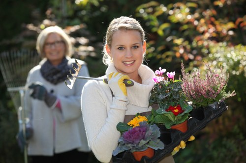 A young woman with blonde hair, wearing a cream knitted sweater and yellow gardening gloves, is smiling at the camera while holding a black tray filled with various potted flowering plants, including purple, yellow, and red blooms, in a well-maintained garden with lush greenery, shrubs, and trees in the background. Behind her, an older woman with glasses, wearing a light jacket and scarf, is also smiling and holding a garden fork, indicating outdoor gardening activity. The garden features a mix of grass, flowering plants, and foliage, with natural sunlight illuminating the scene, suggesting a clear, bright day in Ickenham, suitable for gardening services such as planting and lawn care by Lawn Mowing Ickenham.